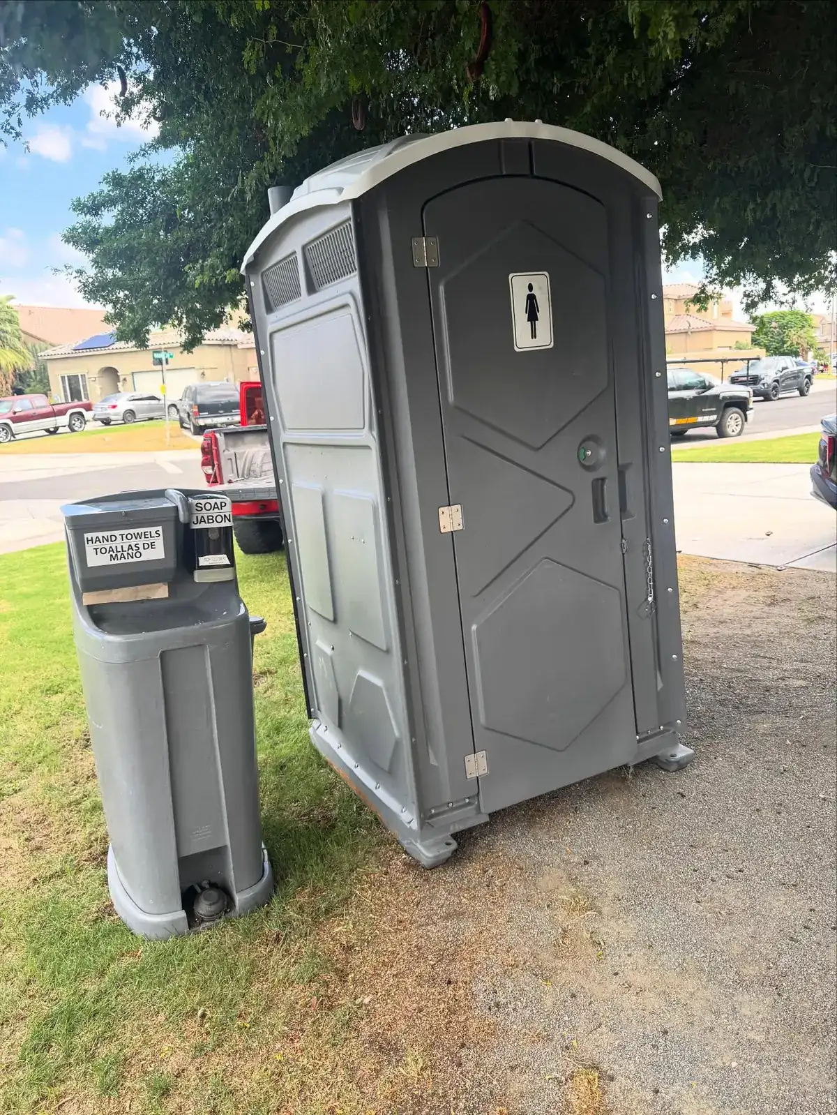 Grey standard porta potty with a hand-washing station beside it